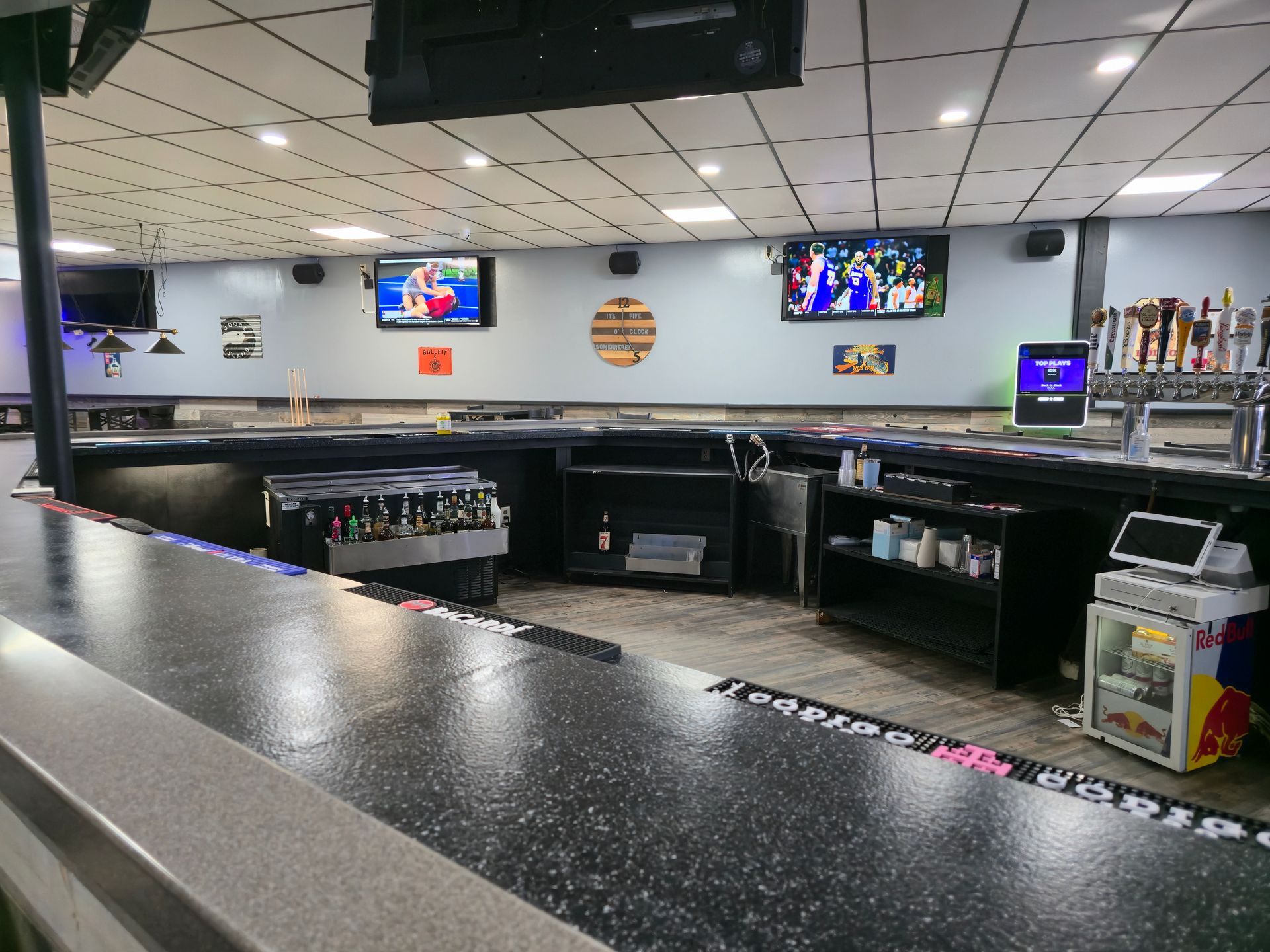 A view of a modern, empty bar with dark countertops, multiple TVs, beer taps, and a Red Bull refrigerator.