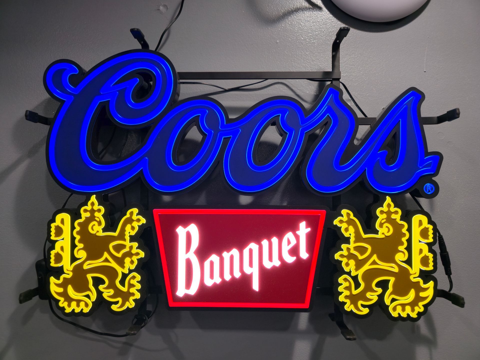 A neon Coors Banquet sign with blue cursive lettering, a central red trapezoid, and flanking gold lion illustrations.