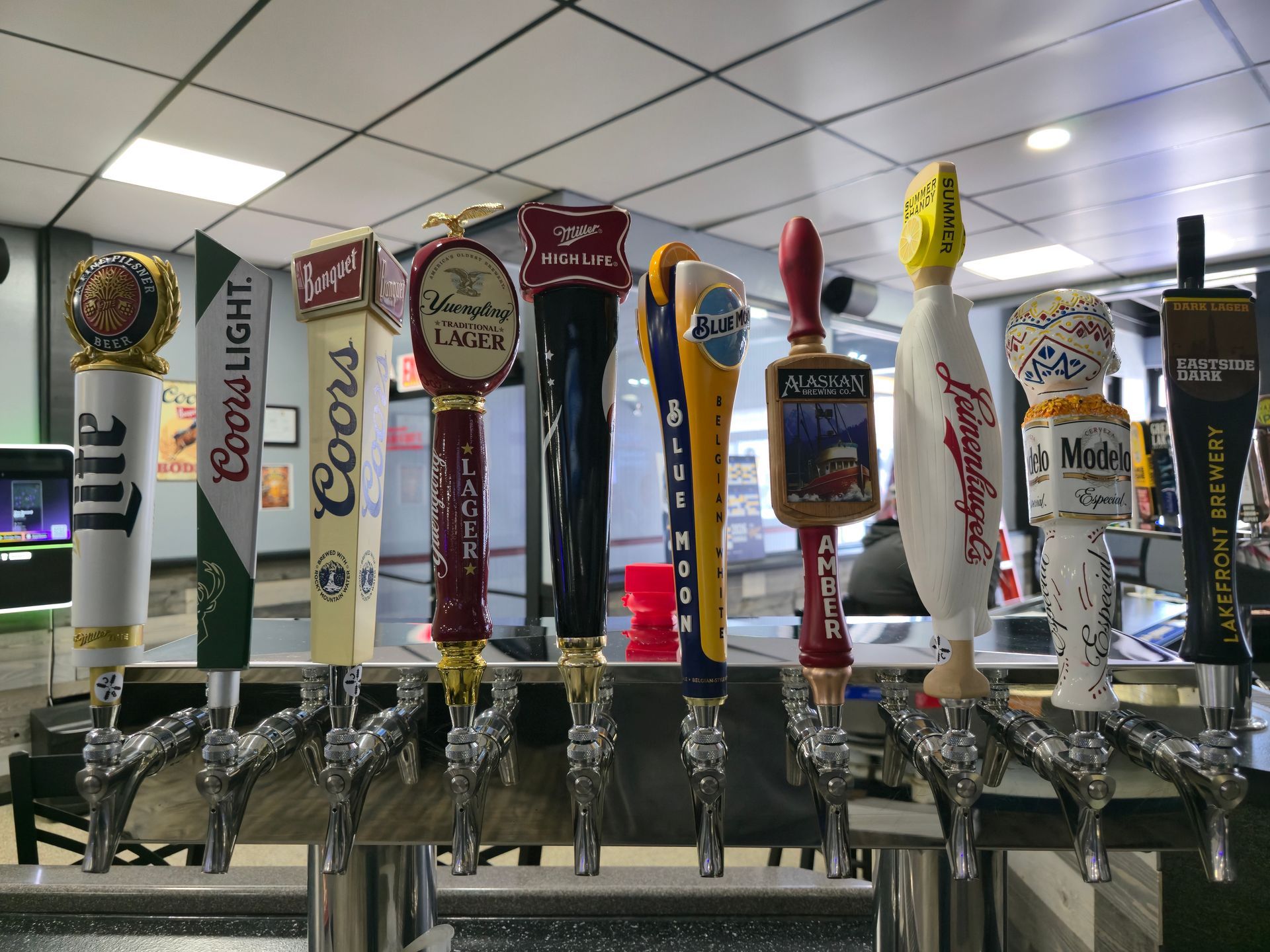 A row of beer tap handles from various brands arranged on a metal bar in a brightly lit indoor venue.