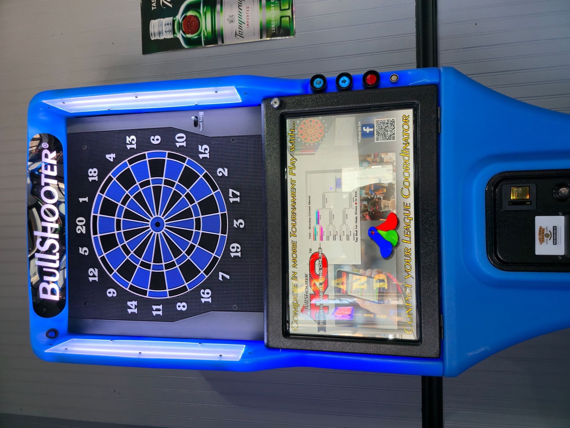 A blue BullShooter electronic dartboard with a side touchscreen interface mounted on a gray wall.