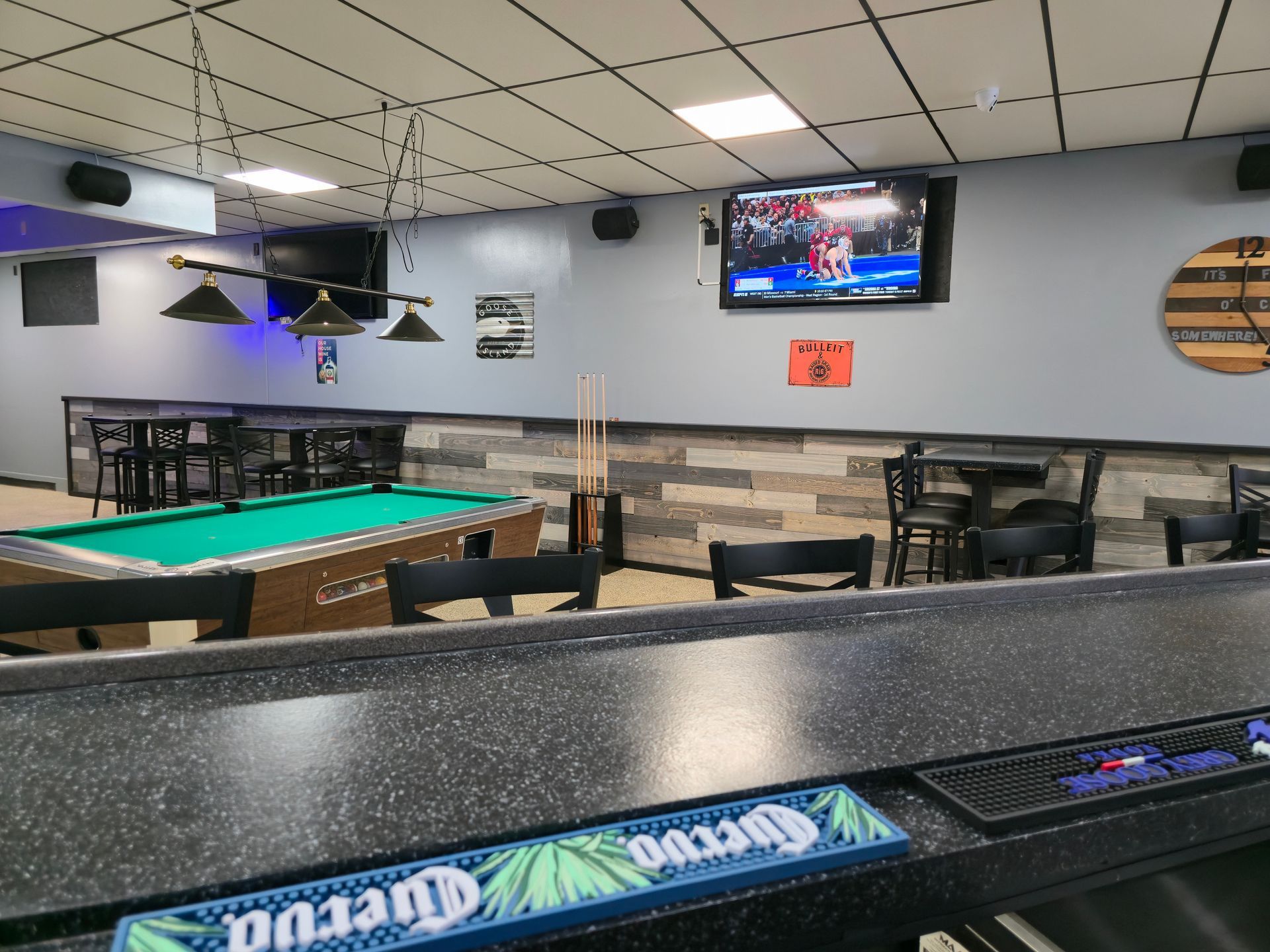 A pool table with green felt in a sports bar, viewed from behind a bar counter with a Cuervo branding mat.