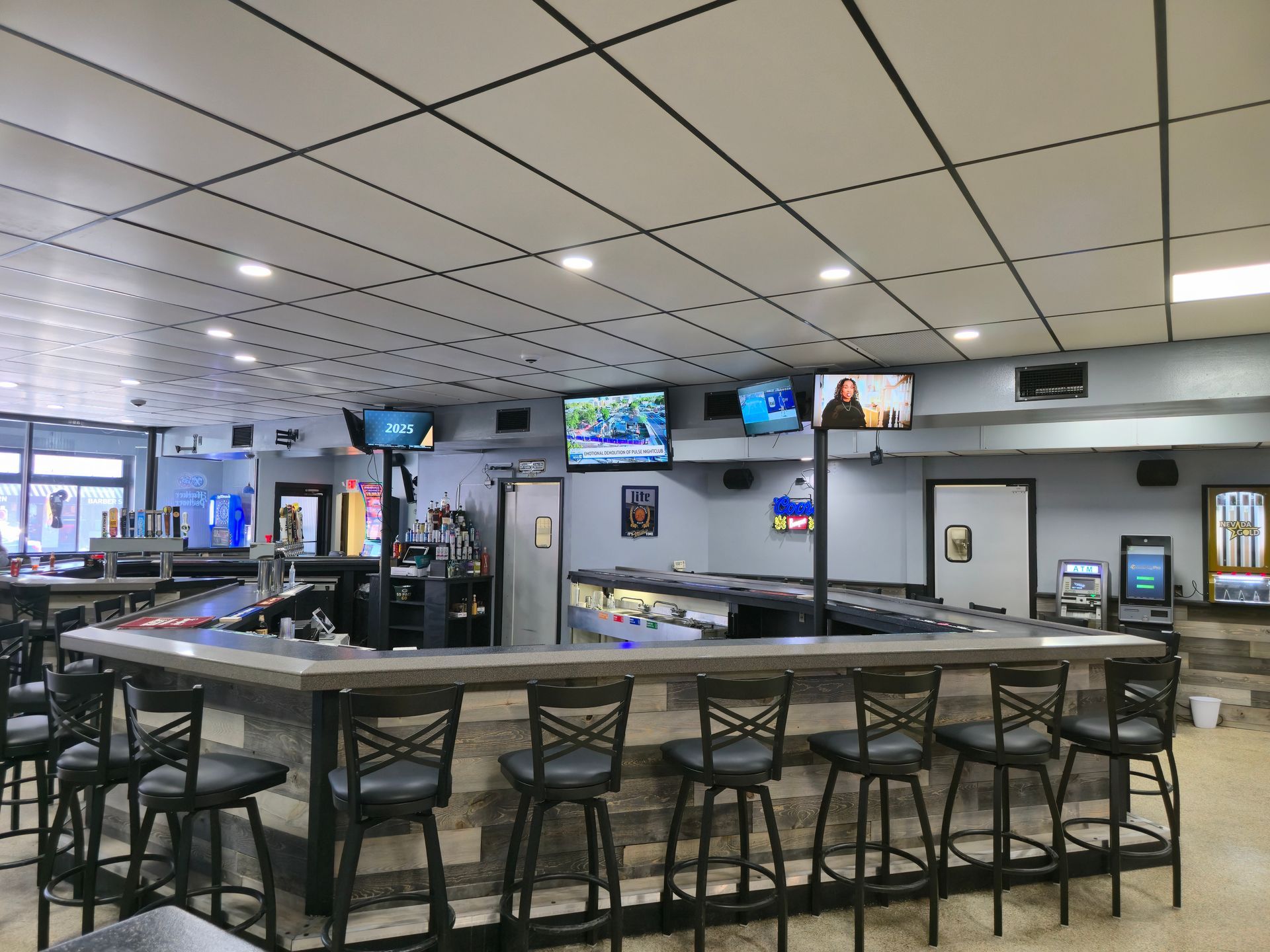 An empty bar featuring a U-shaped counter with several black stools, ceiling tiles, and multiple mounted televisions.