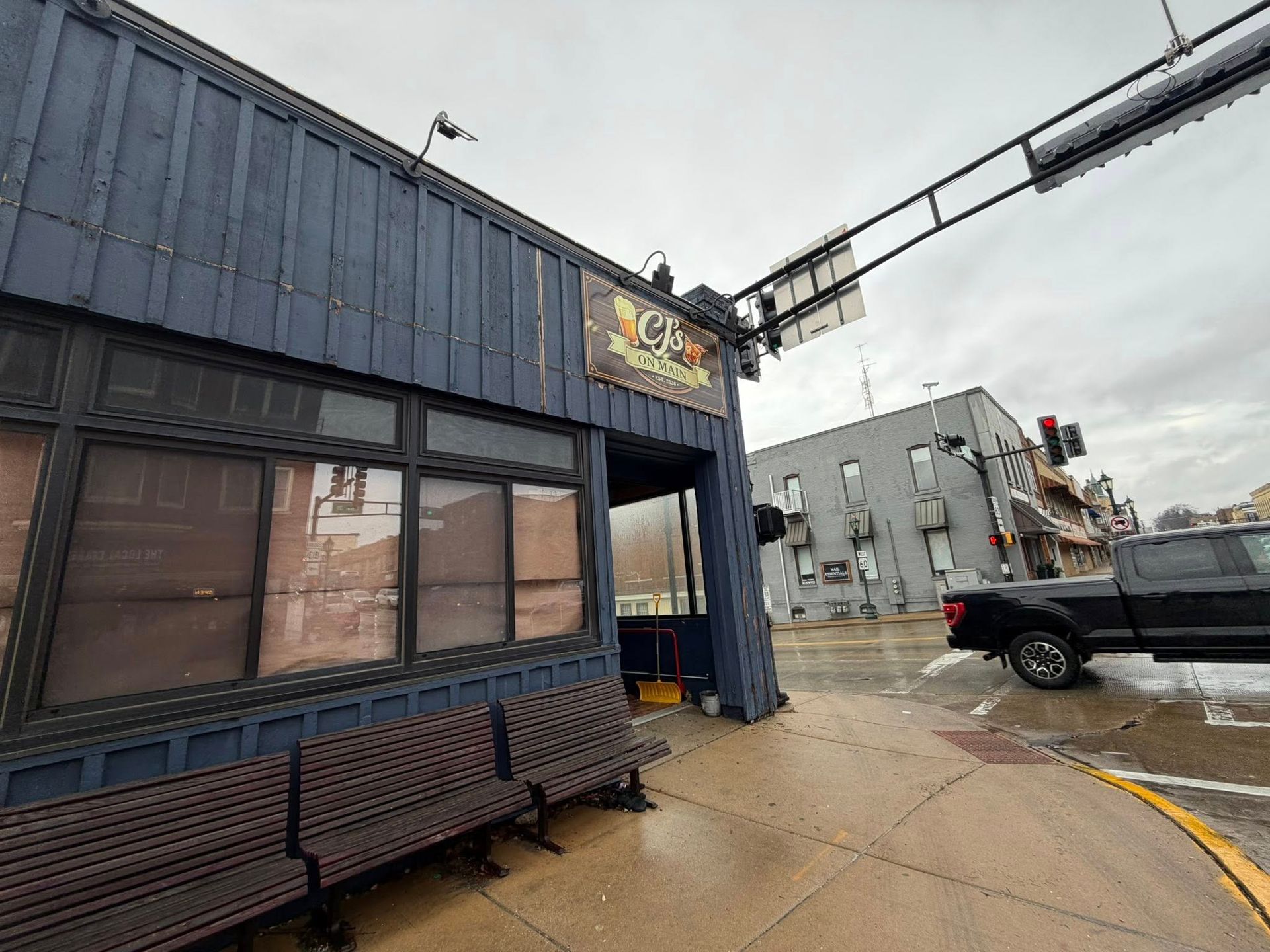A dark blue storefront with a weathered wooden bench on a sidewalk, next to a street intersection and a grey building.