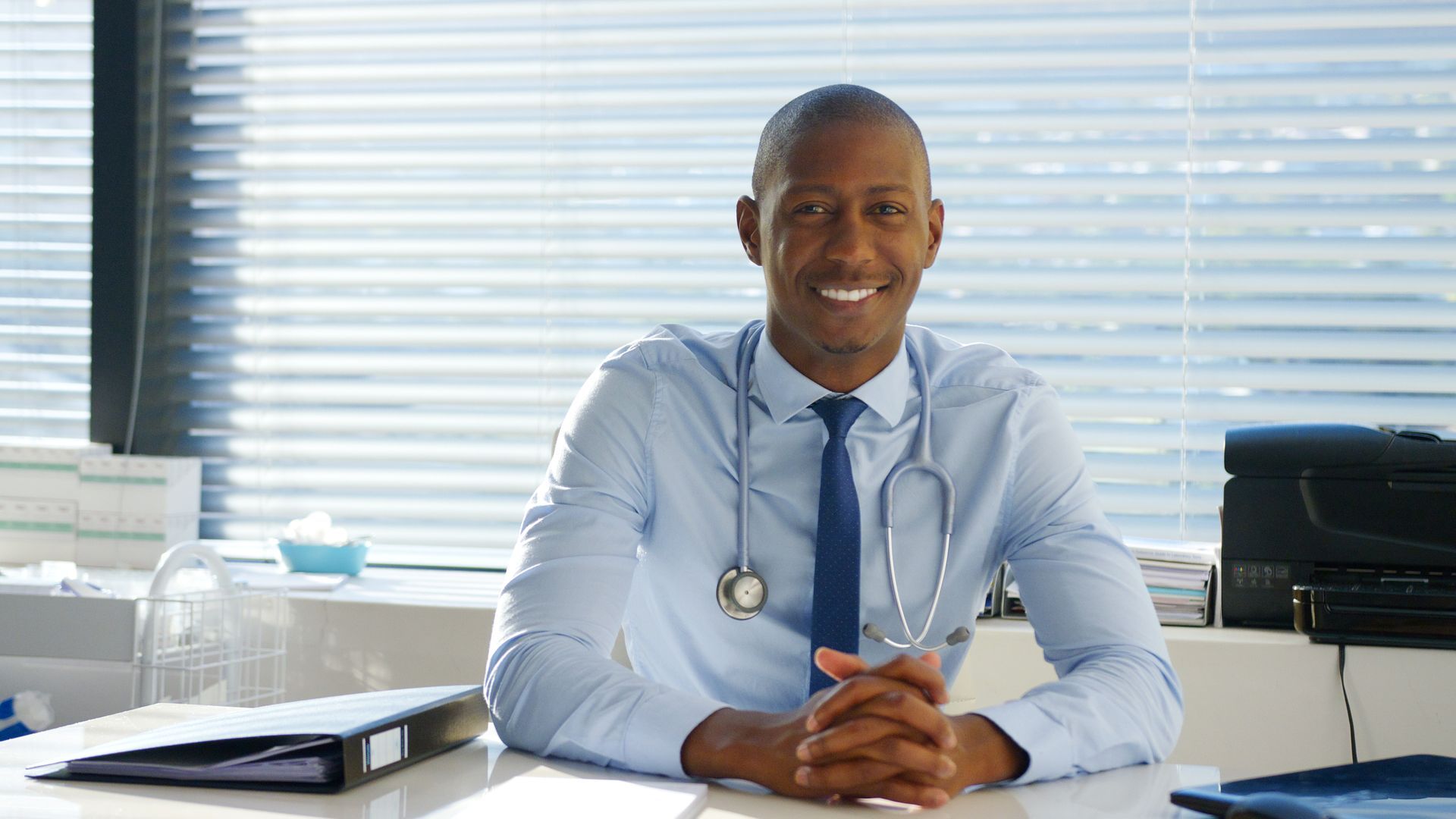 A doctor is sitting at a desk with his hands folded and smiling.