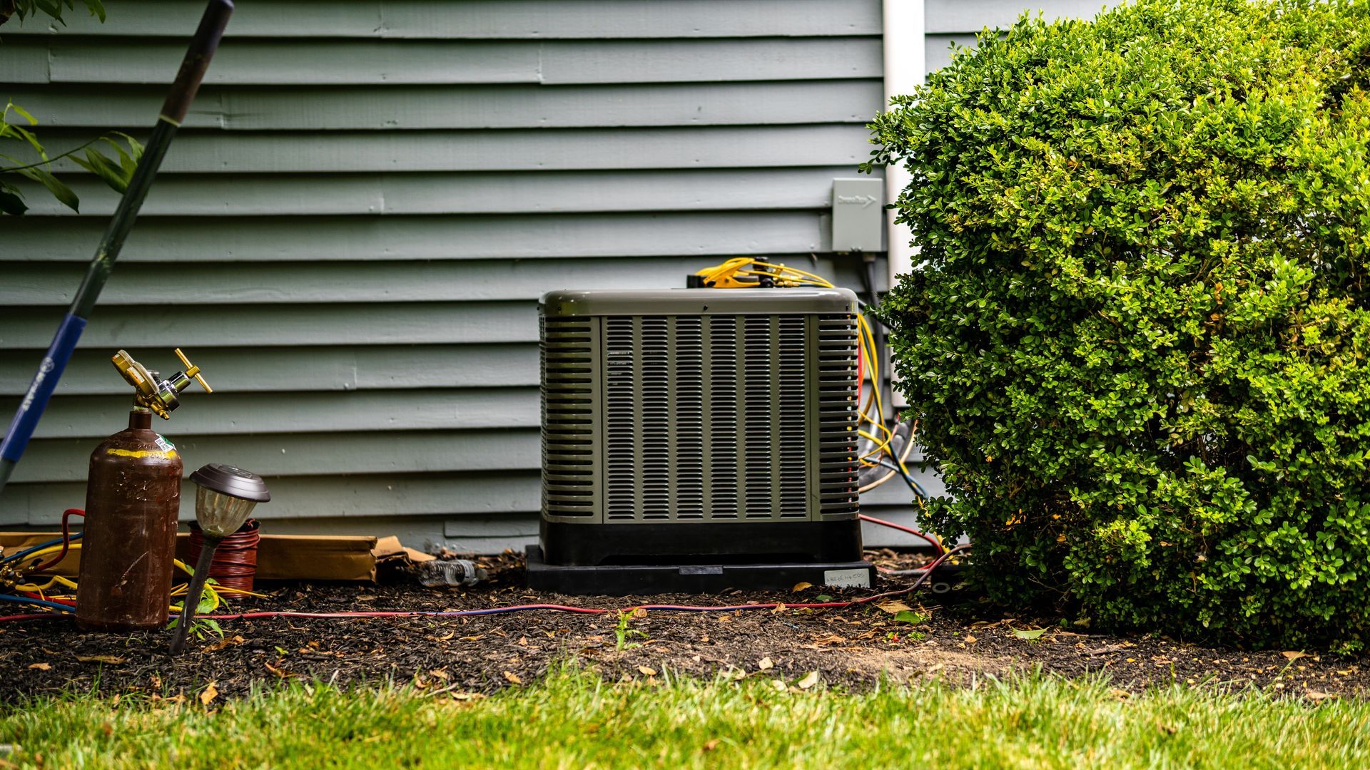 An air conditioner is sitting on the side of a house next to a bush.