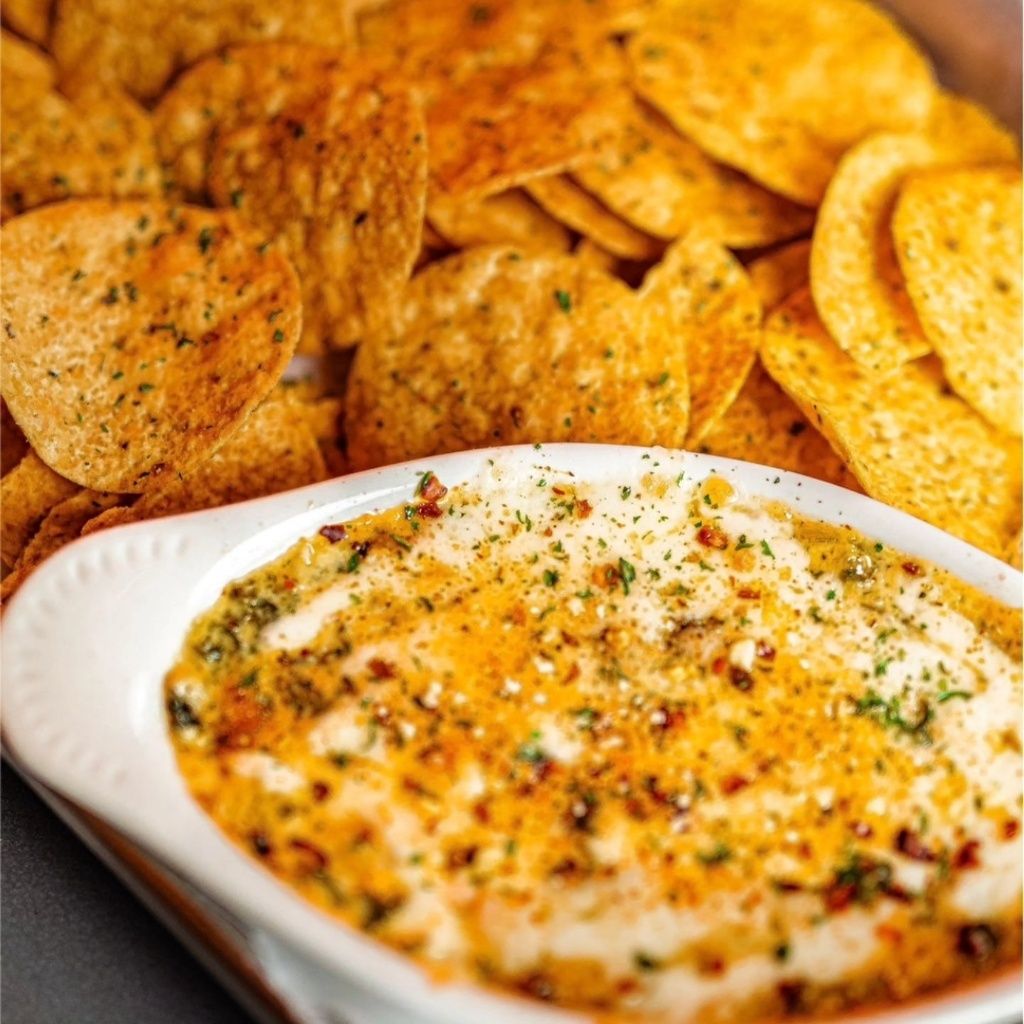 A white bowl of warm, cheesy spinach dip garnished with herbs and red pepper flakes, served with a side of tortilla chips.