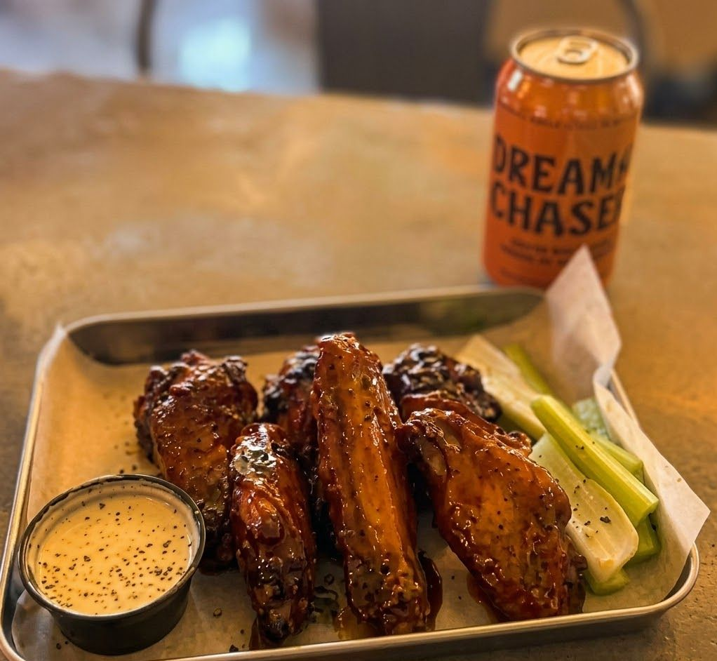 A serving of glazed chicken wings with celery sticks and a side of dip next to a can of Dream Chaser beer.