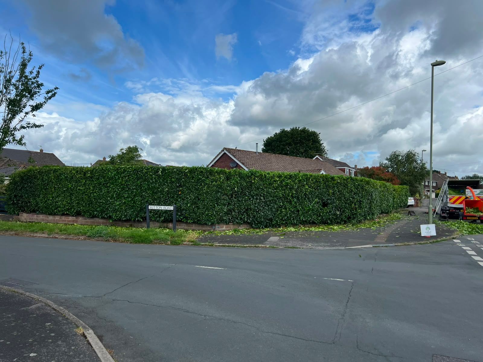 Quiet suburban street corner with a tall hedge, red-roofed house, cloudy sky, and an orange excavator in the distance