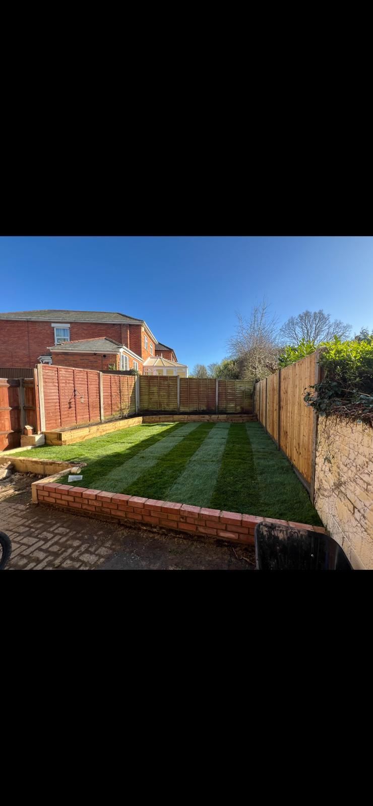 Backyard with a small lawn, brick retaining walls, and a wooden fence under a clear blue sky