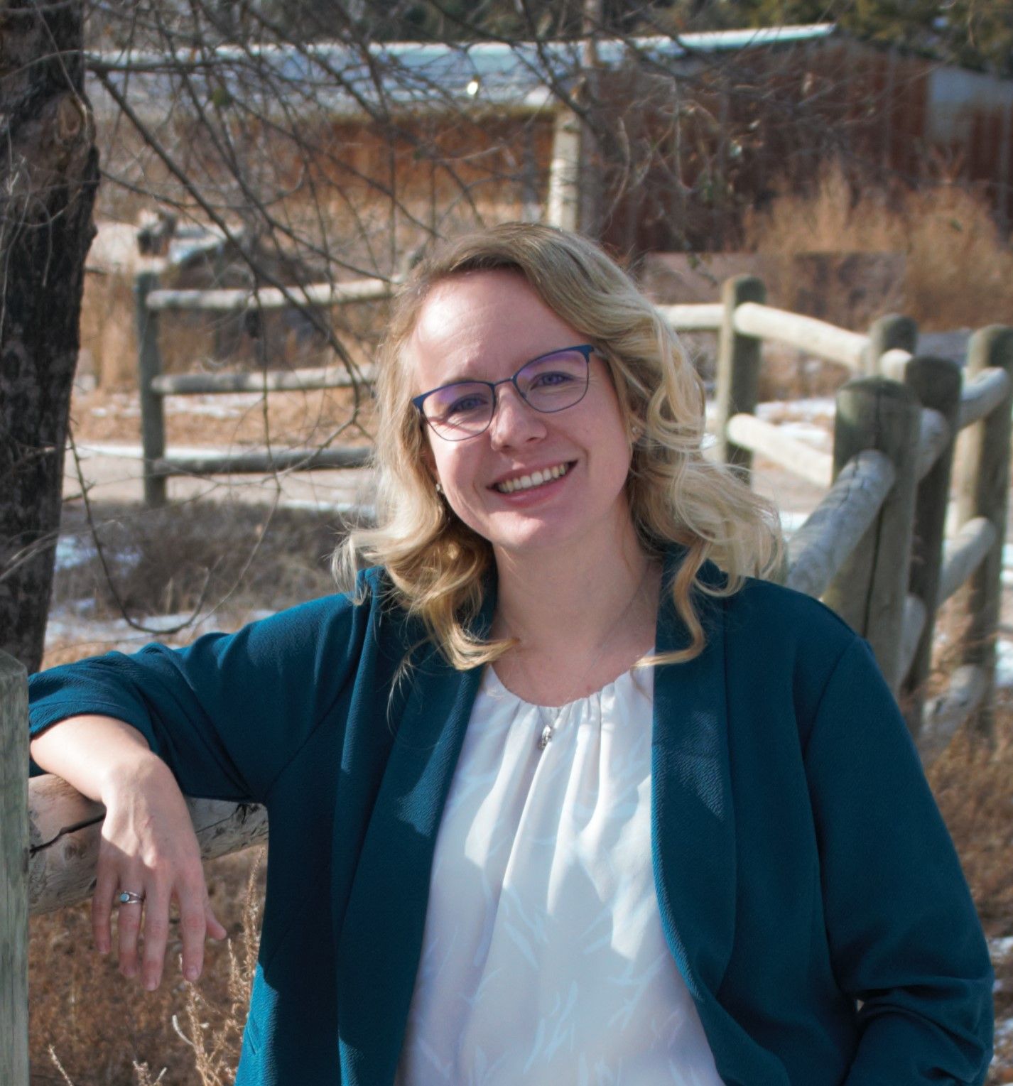 A woman wearing glasses and a blue jacket is leaning on a wooden fence.