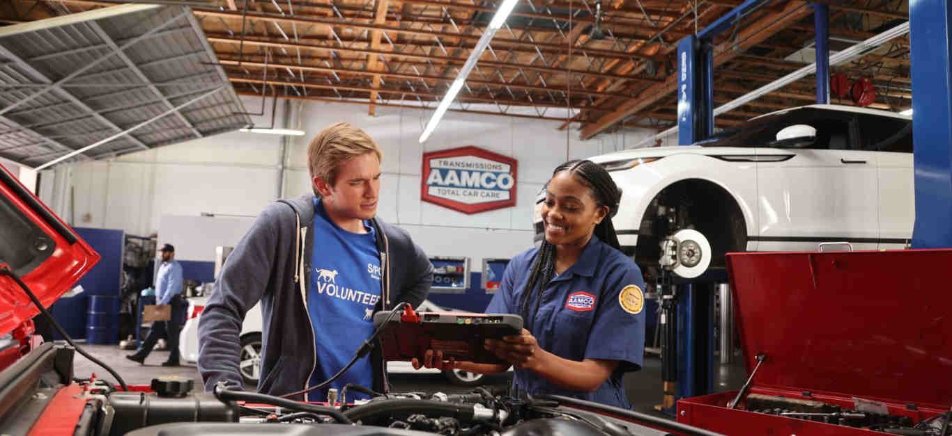 An AAMCO mechanic and customer discuss repairs in a car repair shop. An AAMCO sign is visible.
