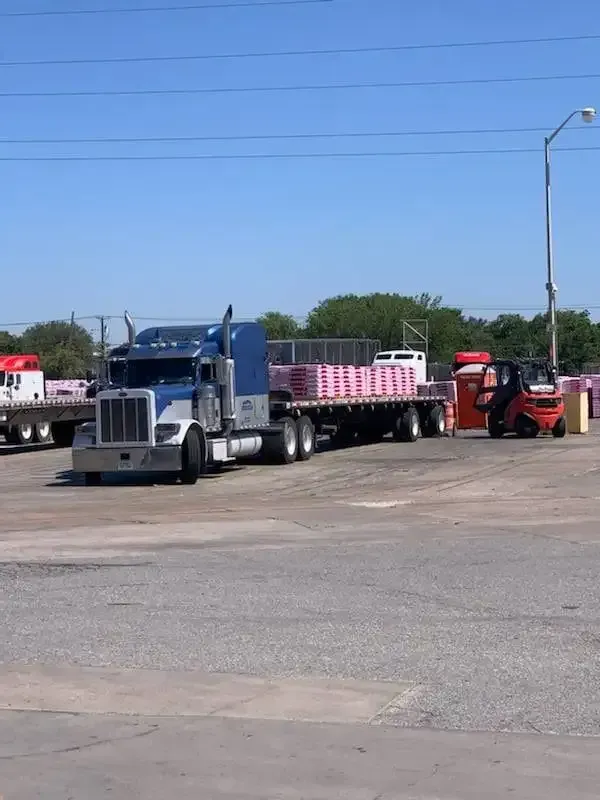 A semi truck with a flatbed trailer is parked in a parking lot next to a forklift.