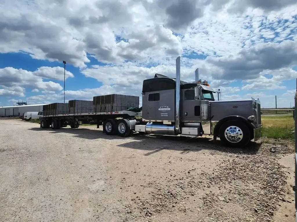 A semi truck with a flatbed trailer is parked in a gravel lot.