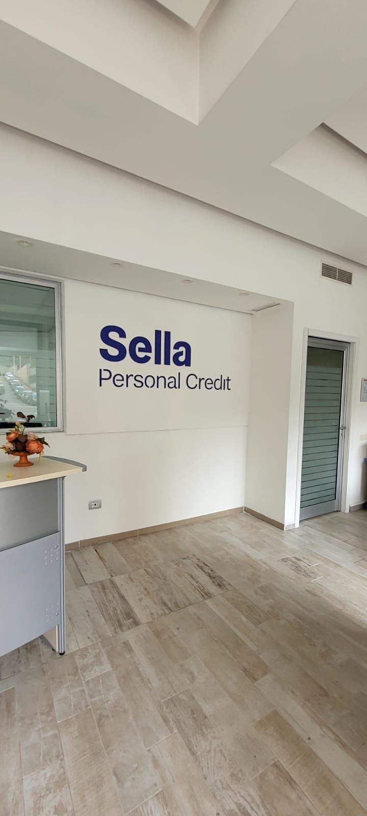 Interno di un ufficio della Sella Personal Credit con pareti bianche, pavimento in legno e porta a vetri.