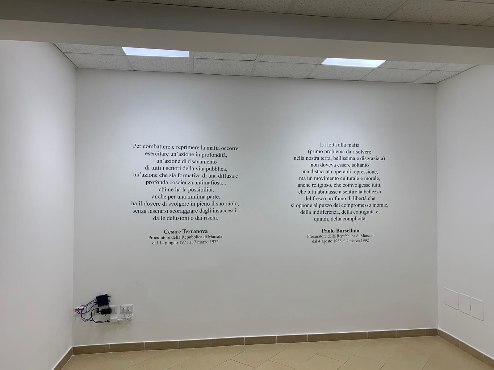 In una stanza con pavimento chiaro, su una parete bianca sono stampate due sezioni di testo nero in spagnolo.