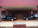 Two black funeral vehicles parked under a covered building entrance with a red roof and brick-colored pillars.