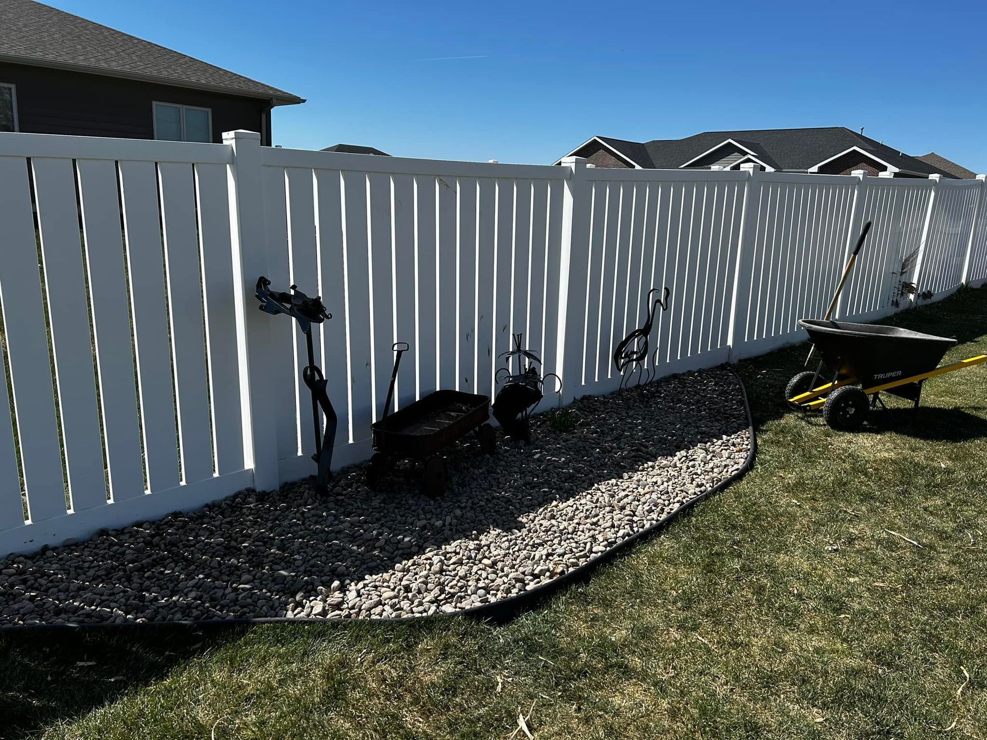 White fence with dark rock border, metal sculptures, and a wheelbarrow on a sunny, grassy lawn.