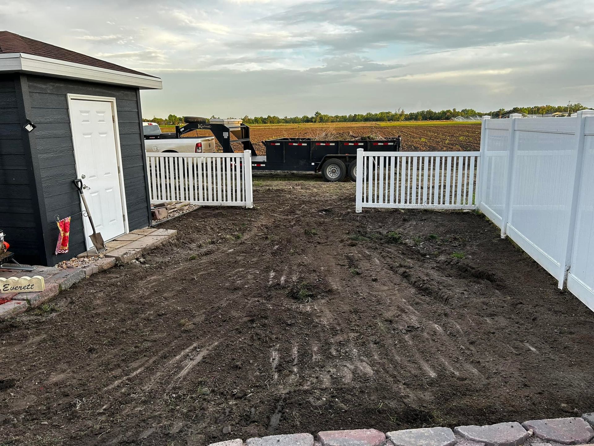 A fenced yard with freshly tilled dirt. A shed, gate, and trailer are in the background under a cloudy sky.