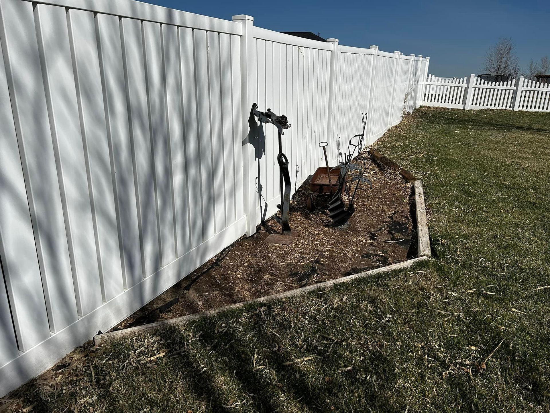 White fence borders a yard with a brown mulch bed and a black faucet fixture.