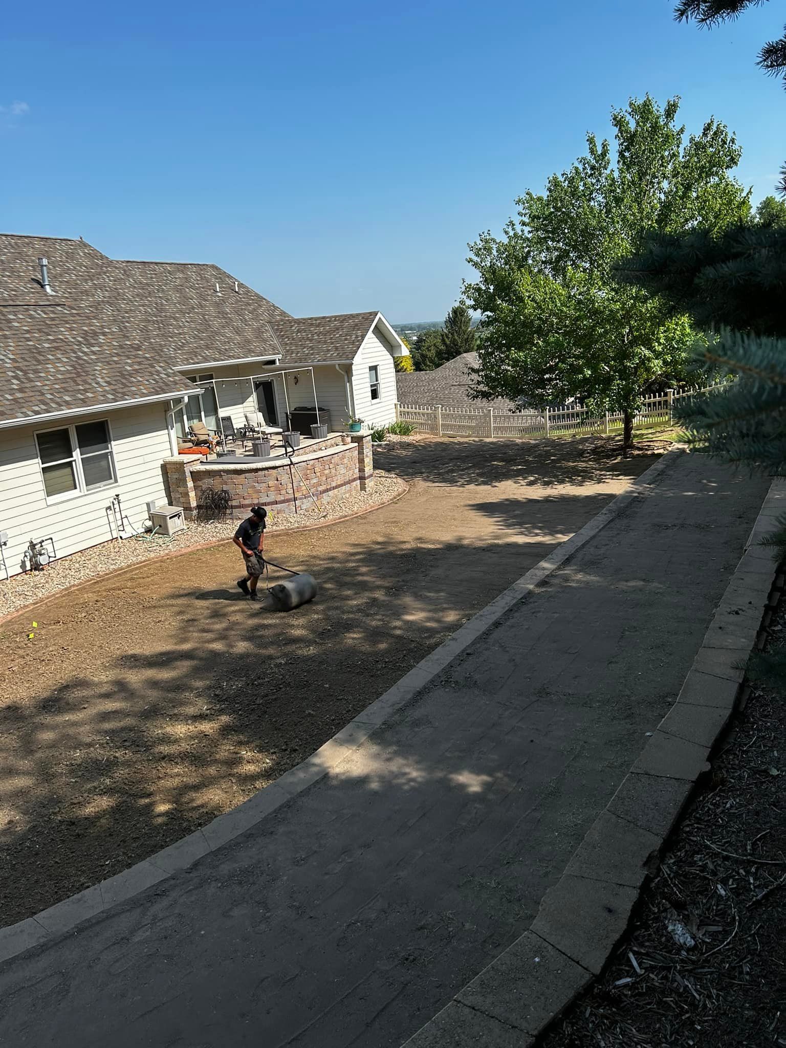 A person mowing a dry lawn near a house and pathway on a sunny day.