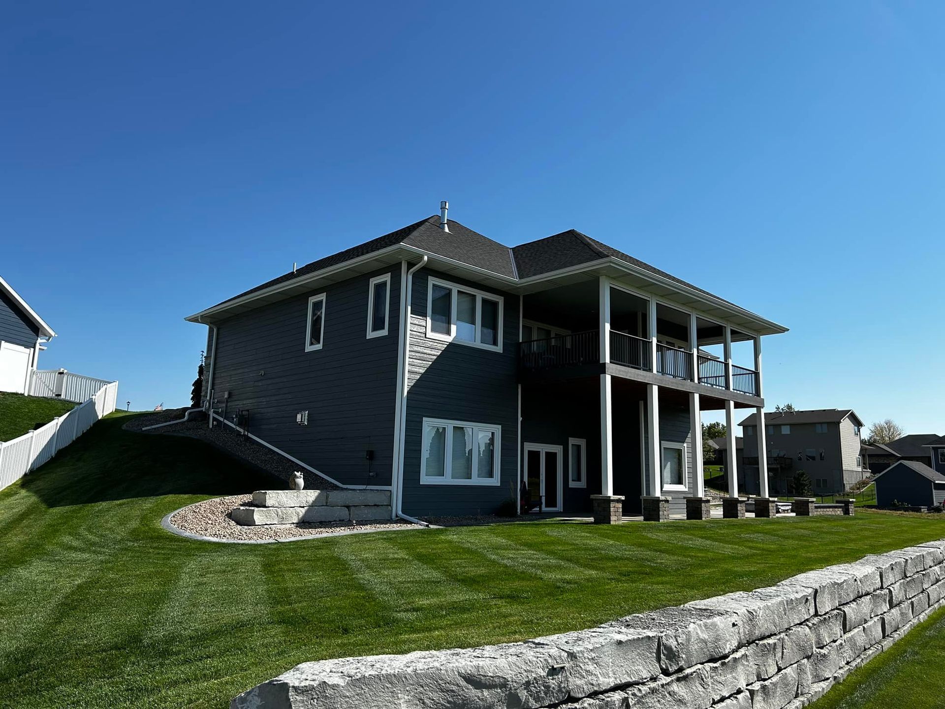 Two-story gray house with white trim, balcony, and lush green lawn under a clear blue sky.
