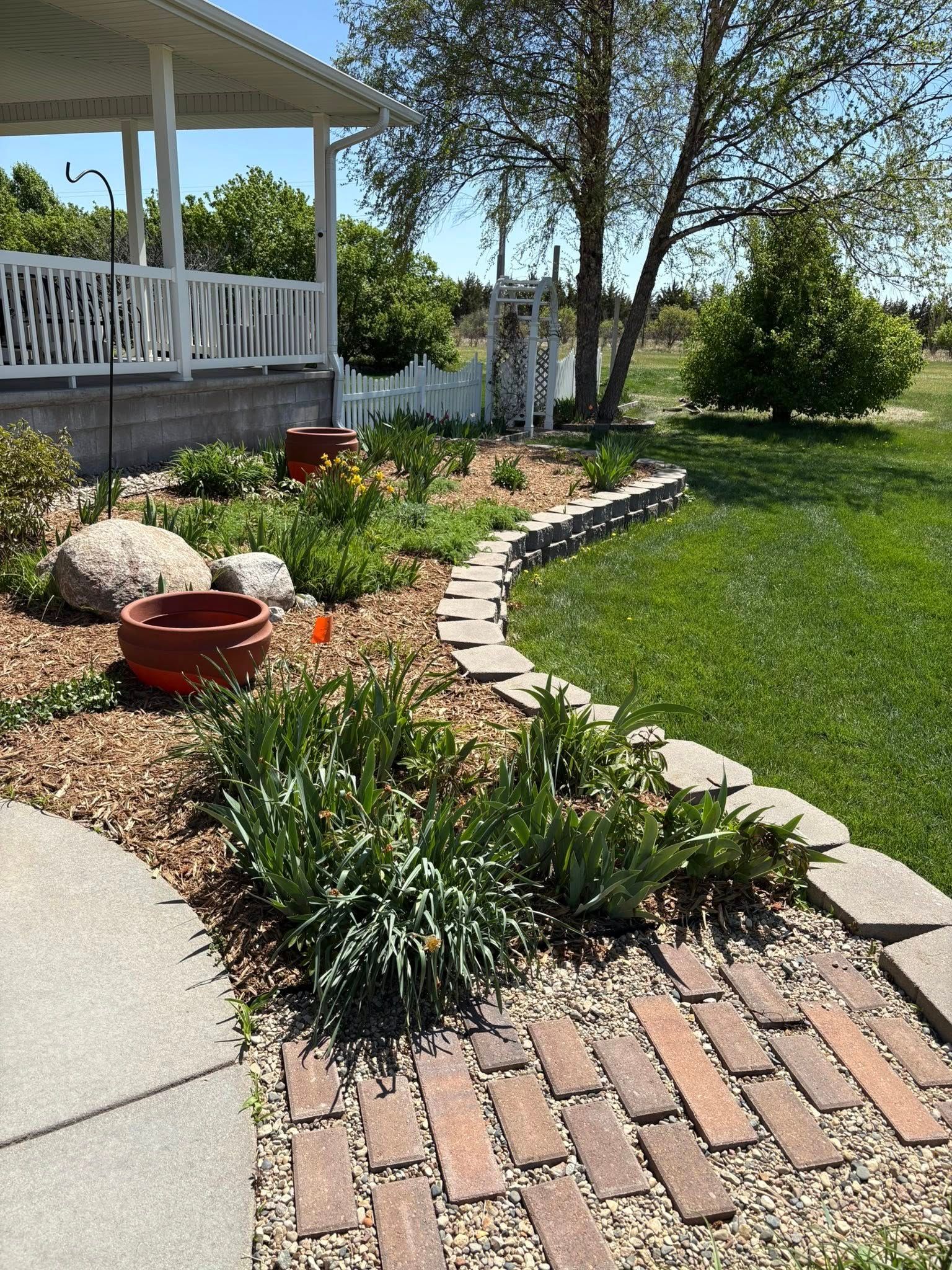 A garden bed with a brick pathway, flowers, and a low stone wall, next to a grassy lawn and a white porch.
