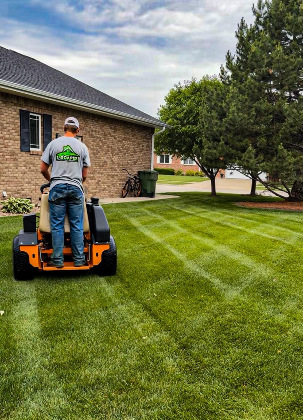 Man mowing lawn with a zero-turn mower; green grass, brick house, sunny day.