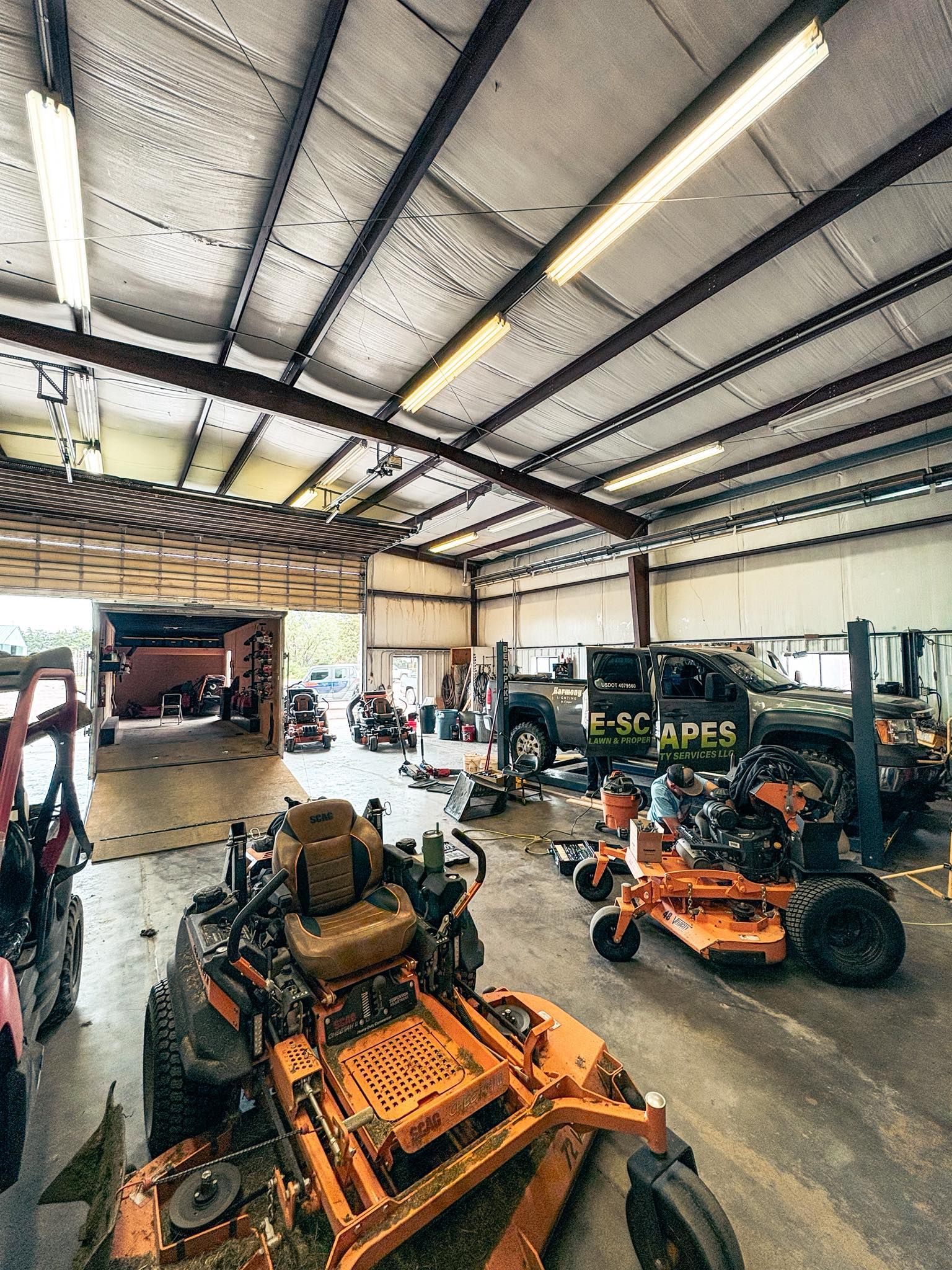 Inside a workshop: orange lawnmowers and a pickup truck; various tools, machinery; overhead lighting.