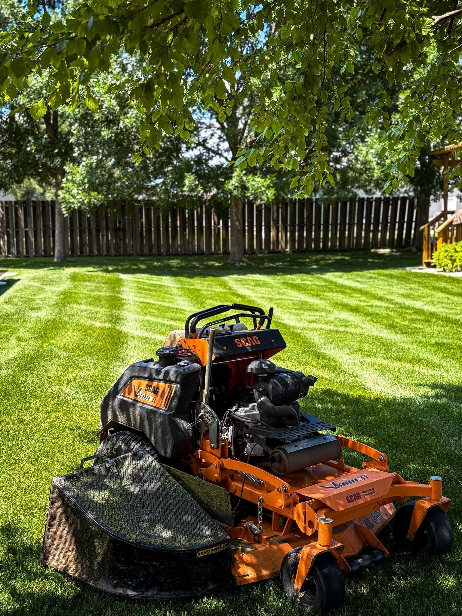 Orange lawnmower on striped green lawn, under trees, in front of wooden fence.
