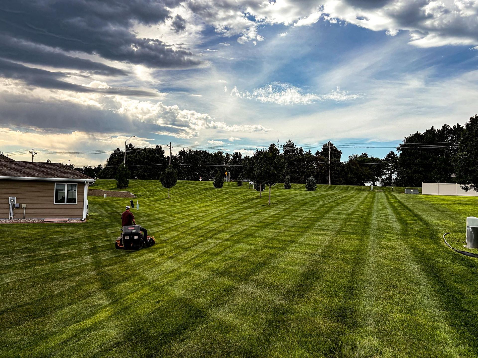 A person mowing a large, striped lawn under a cloudy sky with trees and a house in the background.