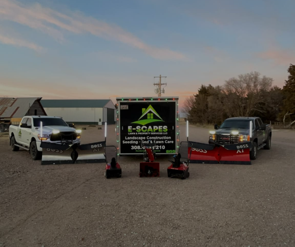 Two trucks with plows and a trailer with landscaping services logo, parked outdoors.