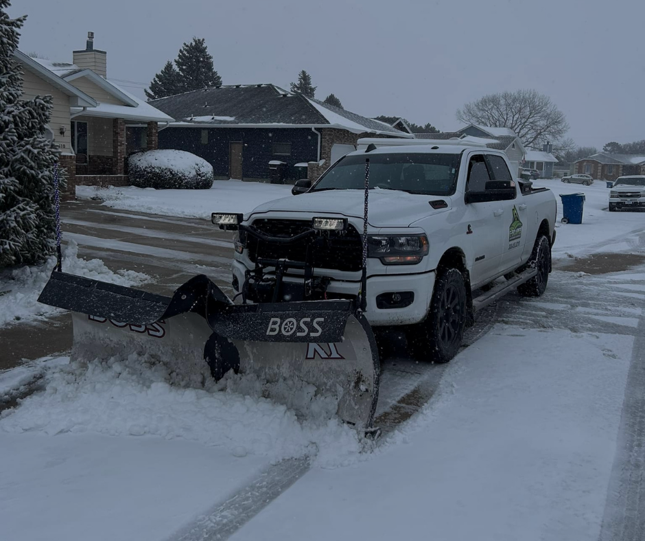 White pickup truck with a snowplow clearing a residential driveway during a snowfall.