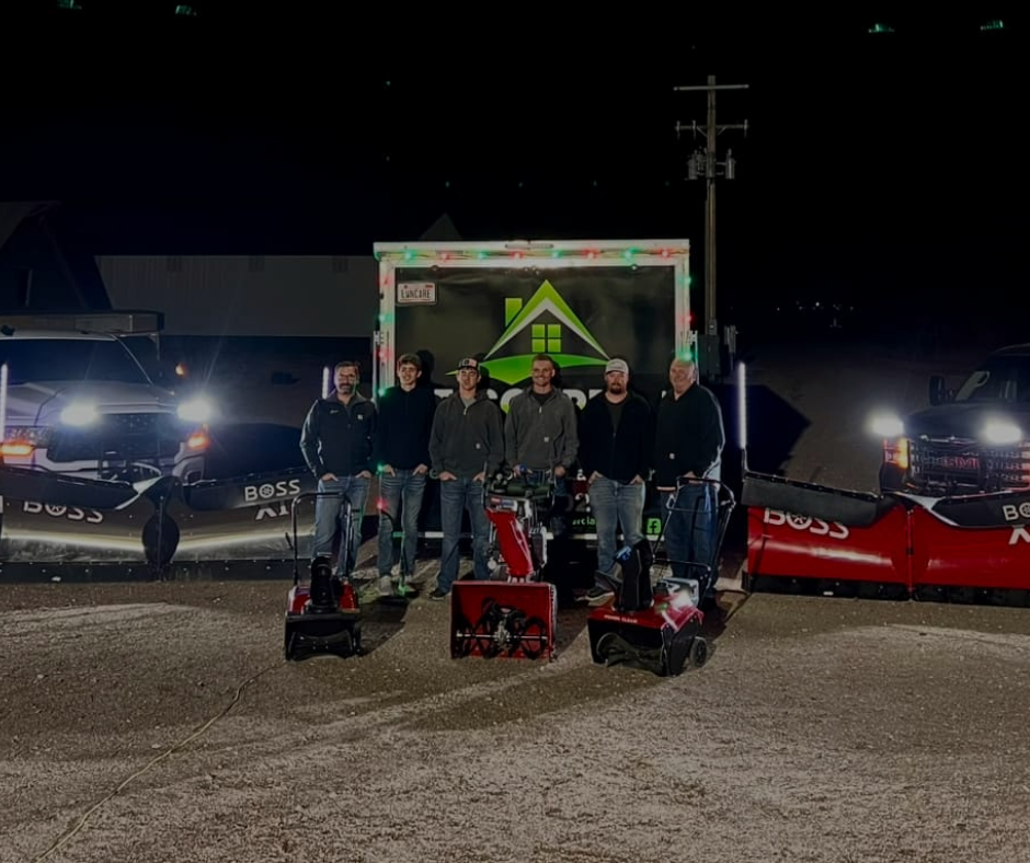 Group of six men with snowblowers posing in front of trucks and a trailer at night.
