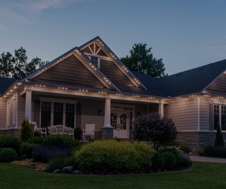 House with white Christmas lights along the roofline at dusk, with a garden in front.