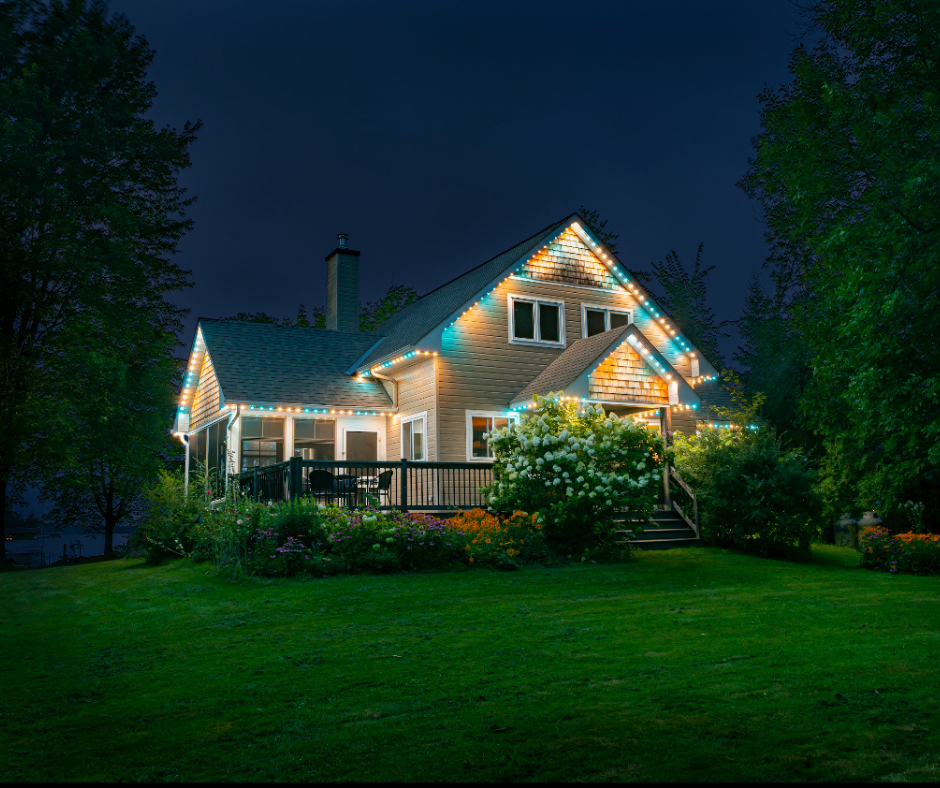 A cozy two-story house at night, decorated with blue and white lights, surrounded by green grass and trees.