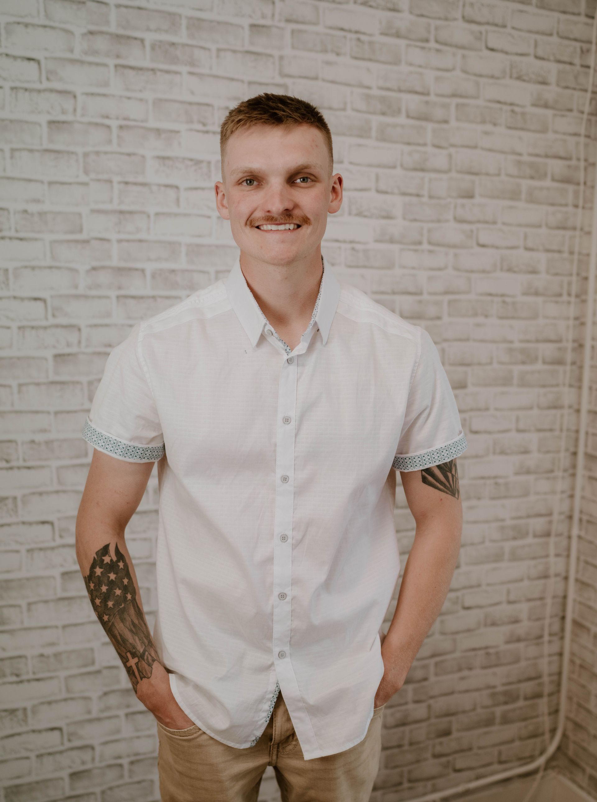 Man in white shirt smiles, hands in pockets, in front of a brick wall. He has tattoos on his arm.