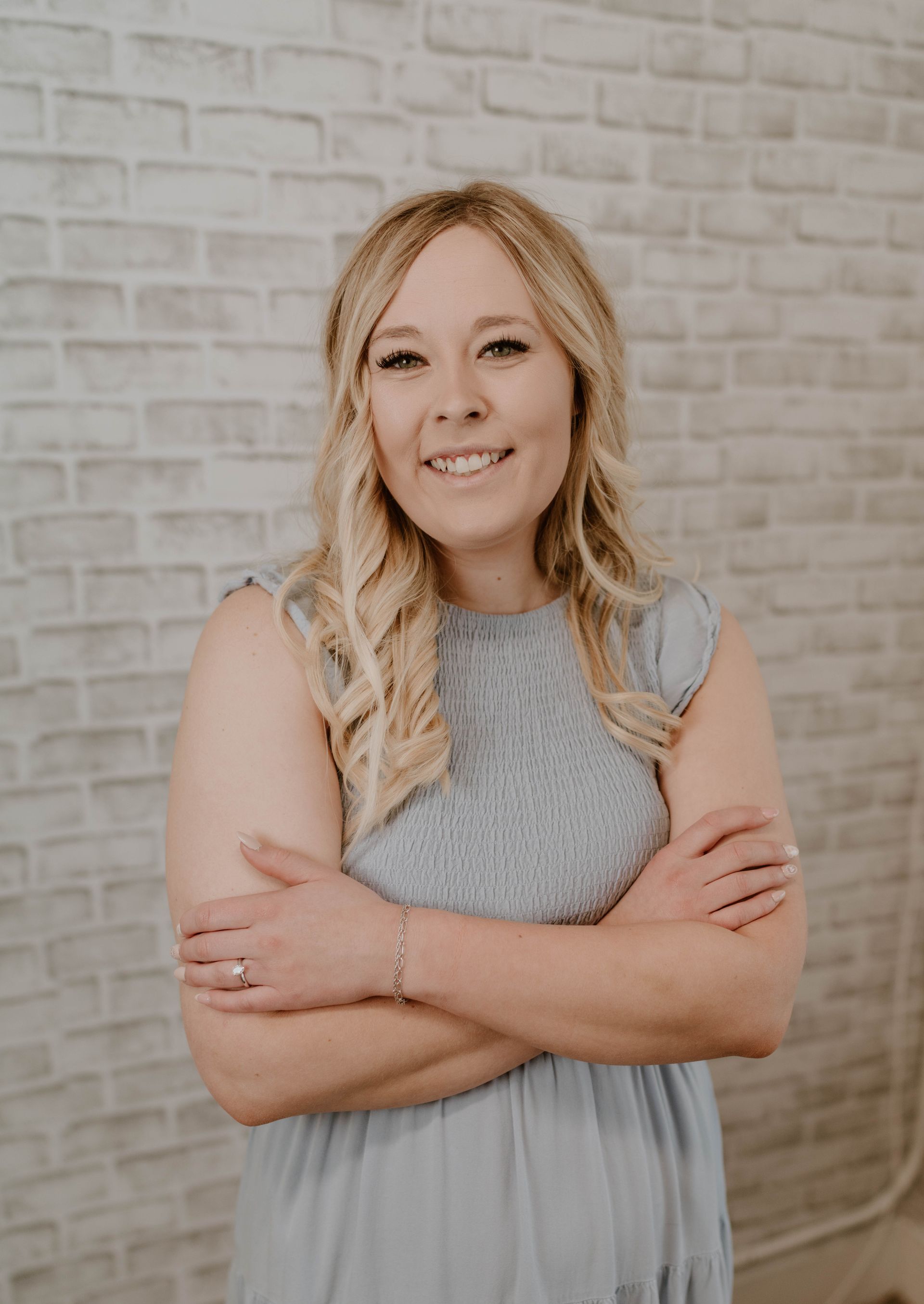 Woman with blonde hair smiles, arms crossed, wearing a gray dress, standing in front of a brick wall.