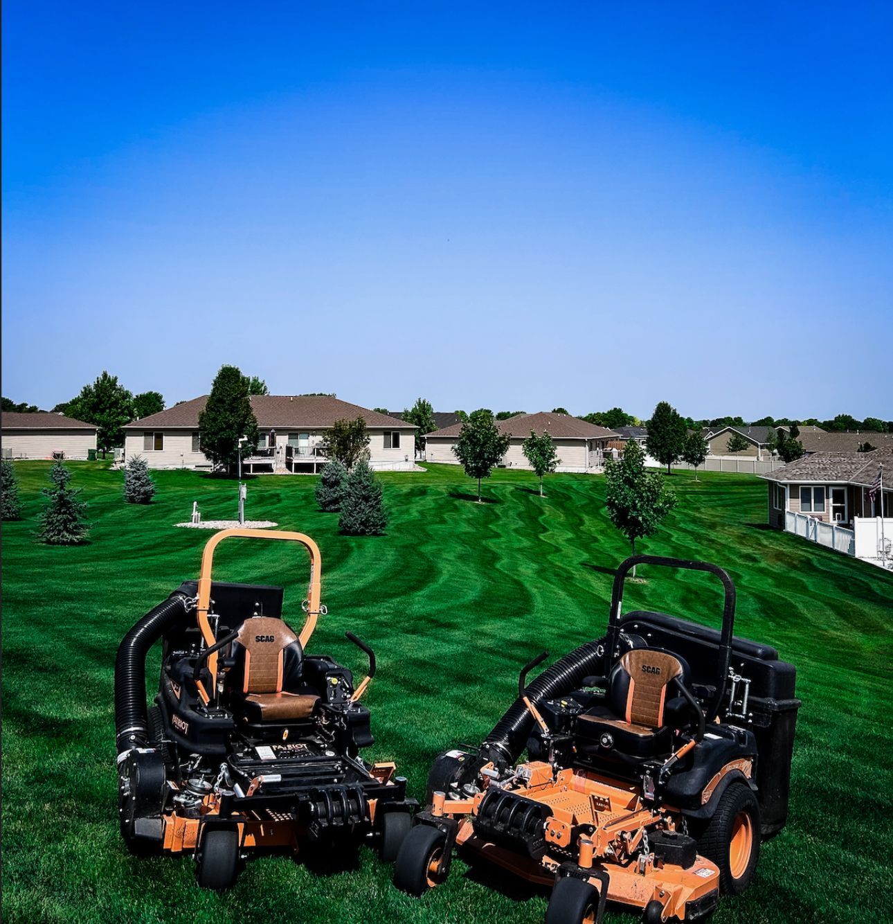 Two orange zero-turn lawnmowers on green grass with houses and a blue sky in the background.