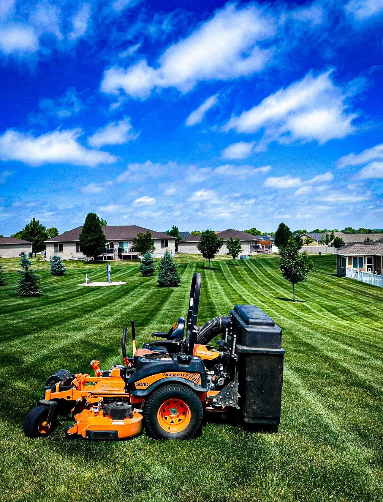 Orange and black lawnmower cutting stripes into a green lawn, with houses and a blue sky.