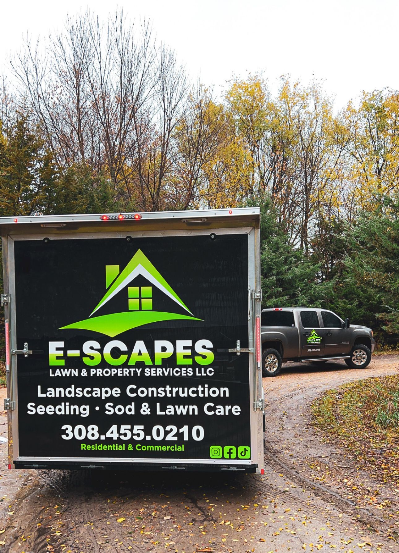 A trailer and truck for E-Scapes landscaping company on a dirt road, with trees in the background.