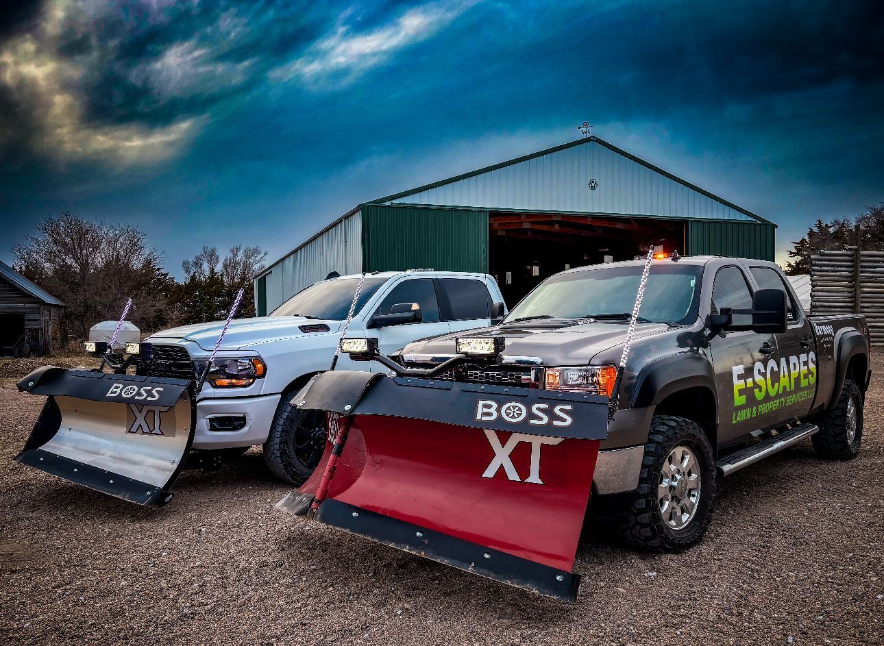 Two trucks with snow plows parked in front of a barn, under a cloudy sky. One plow says BOSS XT.