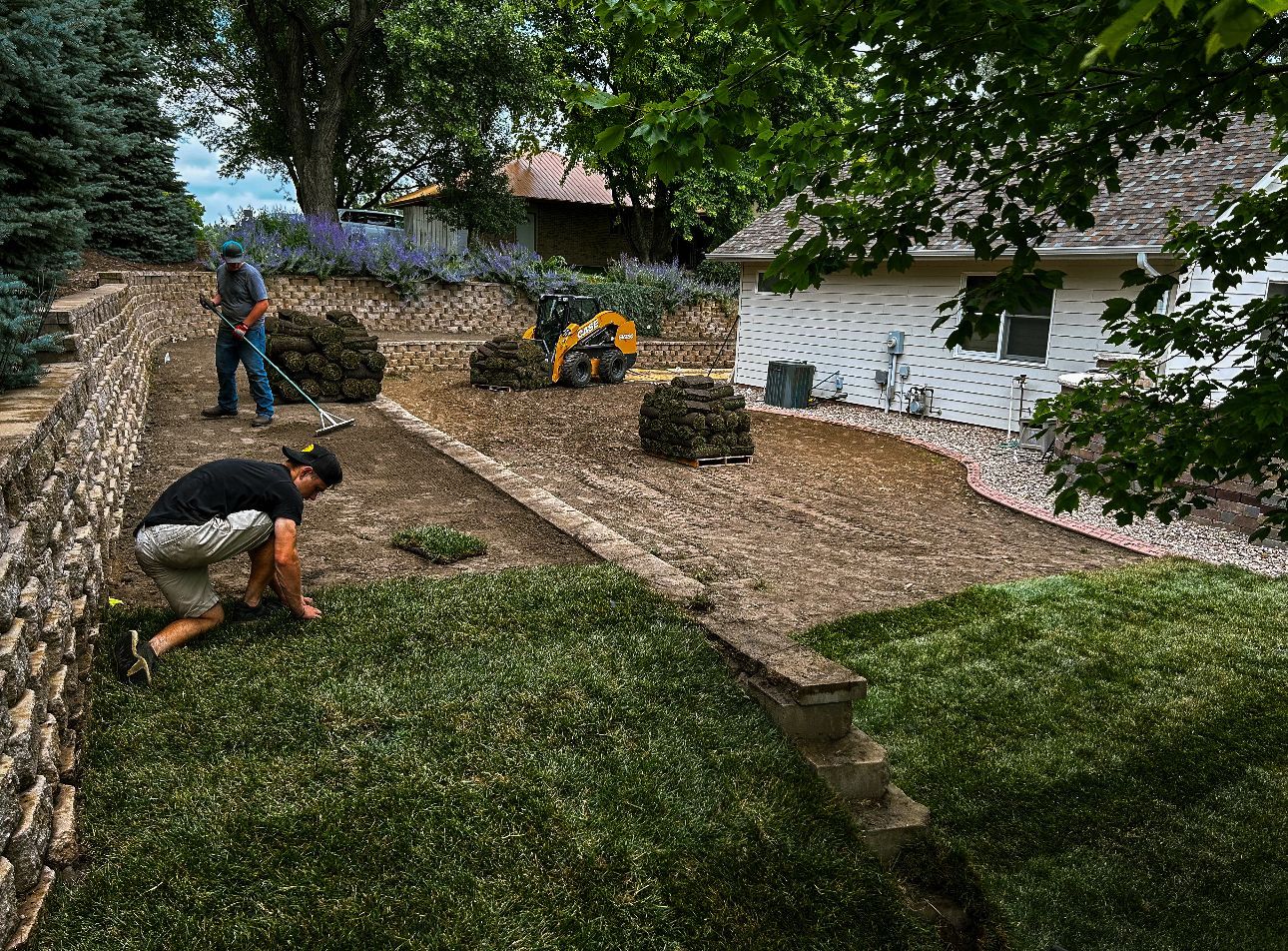Landscapers installing sod on a sloped yard with stone retaining walls and a white house.