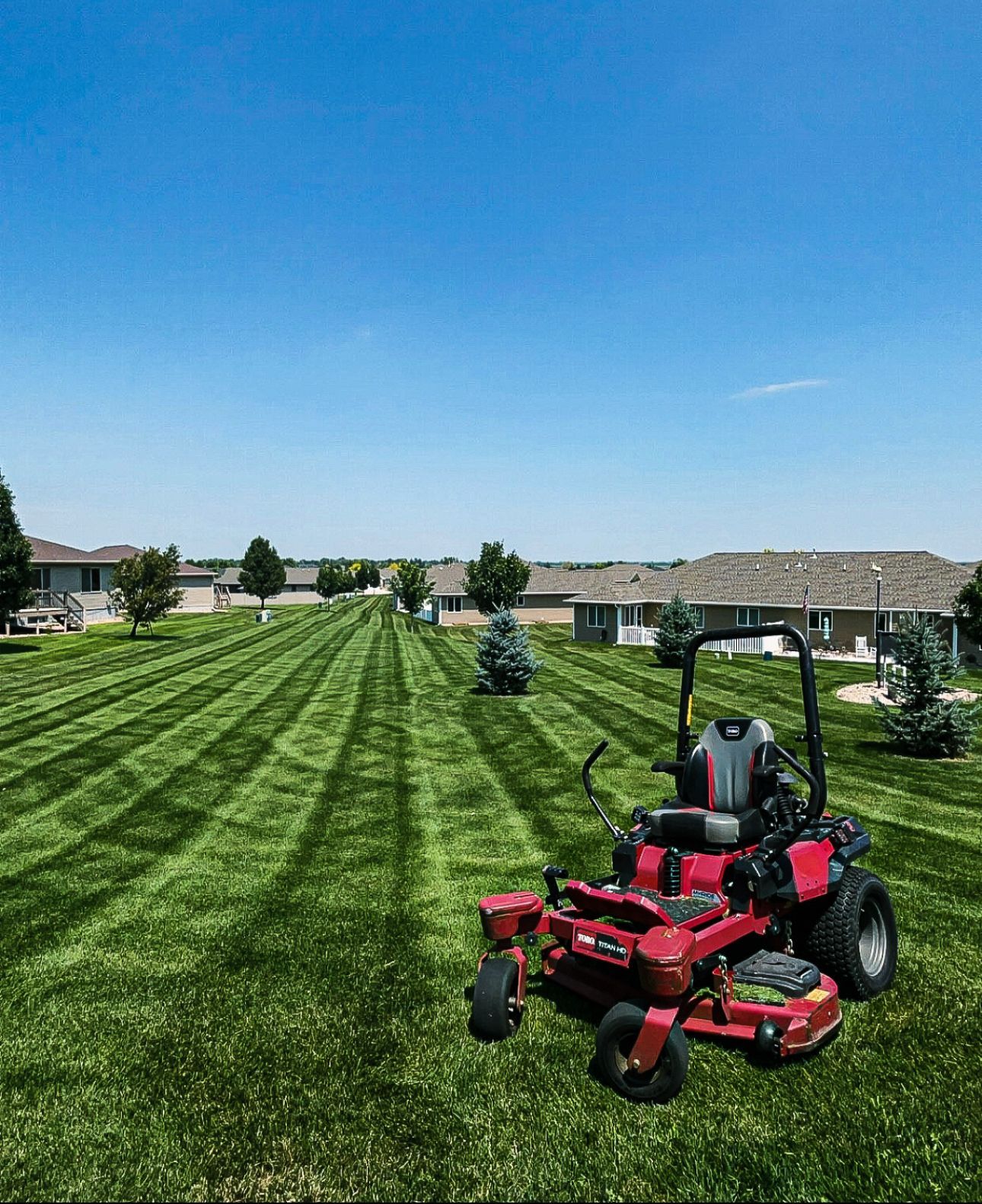 Red zero-turn lawnmower on a striped green lawn under a blue sky. Houses and trees in the background.