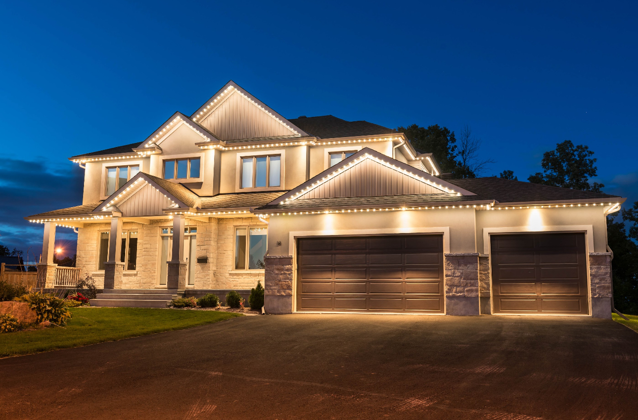 Two-story house at dusk, lit up with outdoor lighting, beige siding, brown garage doors, on a driveway.