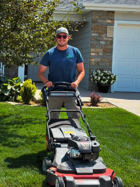 Man mowing a green lawn in front of a house on a sunny day.