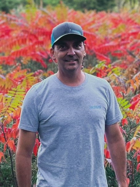 Man in gray shirt and cap smiles in front of red and yellow plants.