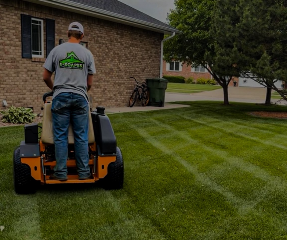 Man mowing a striped lawn with a zero-turn mower in front of a brick house on a sunny day.