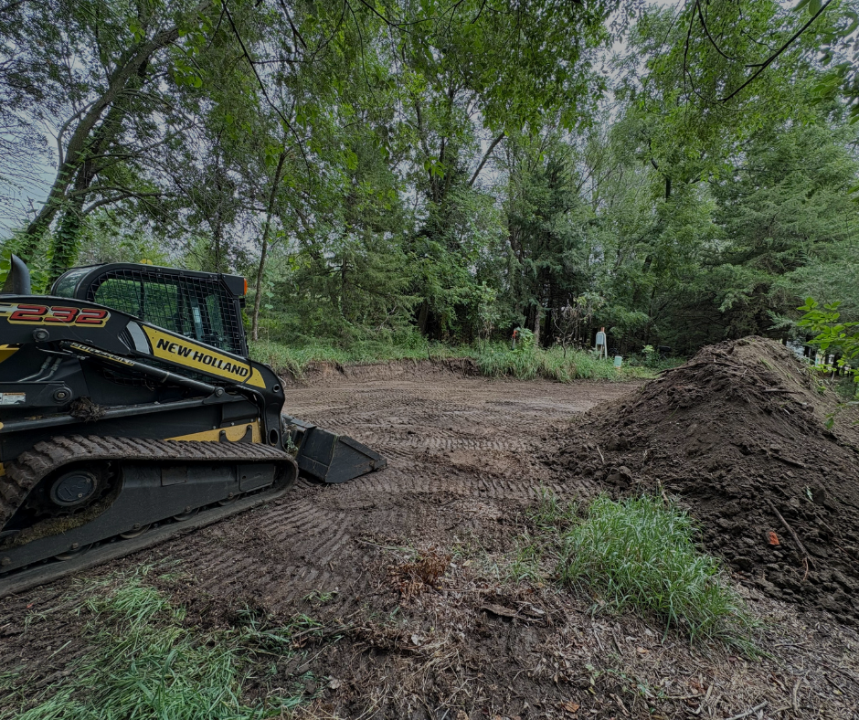 A New Holland skid steer on a dirt plot near trees and a pile of dirt.