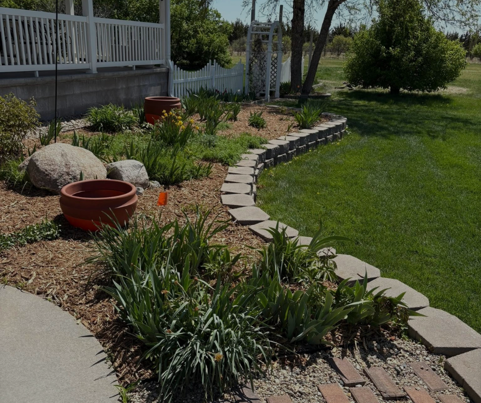 Stone-lined flower bed with greenery, terracotta pots, and mulch, bordering a lawn, with a house in the background.