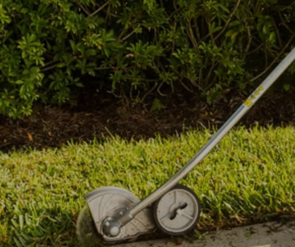 A string trimmer edging along a lawn's edge, with green grass, mulch, and bushes in the background.